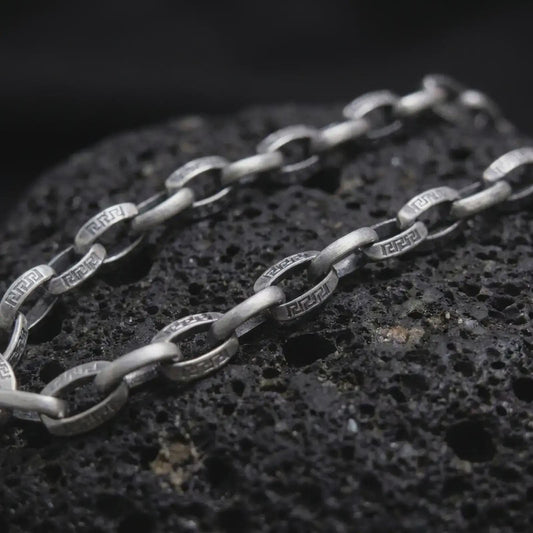 Oxidized Greek key chain necklace rests on dark volcanic rock in a macro studio close-up, showing patterned silver links with ancient-inspired detail.