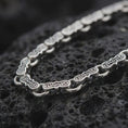 Load image into Gallery viewer, Oxidized Greek key chain necklace rests on dark volcanic rock in a macro studio close-up, showing detailed meander links with ancient-inspired patterning.

