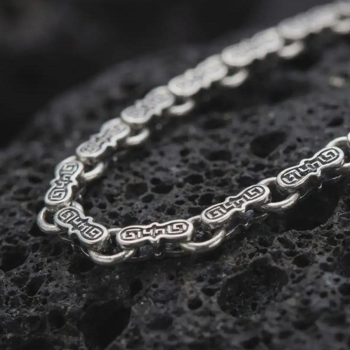 Oxidized Greek key chain necklace rests on dark volcanic rock in a macro studio close-up, showing detailed meander links with ancient-inspired patterning.