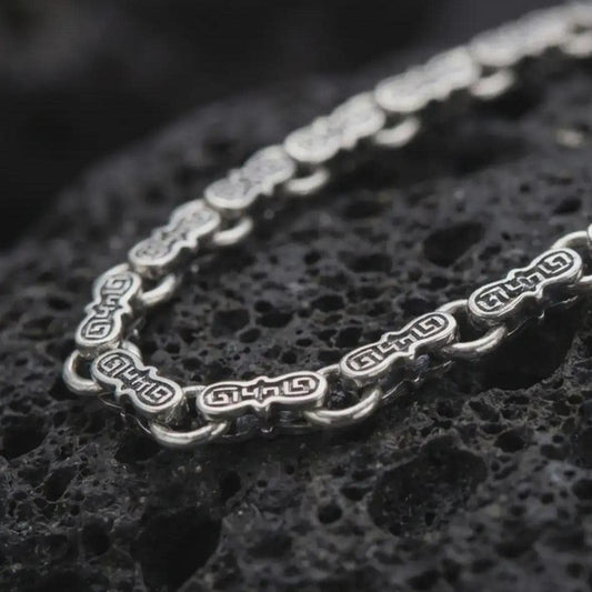 Oxidized Greek key chain necklace rests on dark volcanic rock in a macro studio close-up, showing detailed meander links with ancient-inspired patterning.