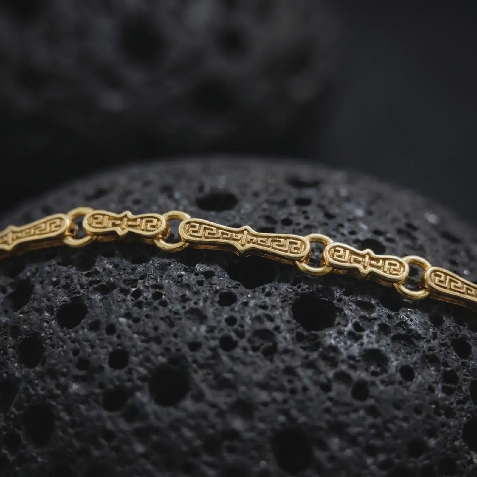 Gold plated Greek key chain necklace sits on textured volcanic rock in a dramatic studio macro shot, highlighting the repeating ancient Greek pattern.