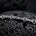 Load image into Gallery viewer, Oxidized Greek key chain necklace rests on dark volcanic rock in a macro studio close-up, showing detailed meander links with antique silver contrast.
