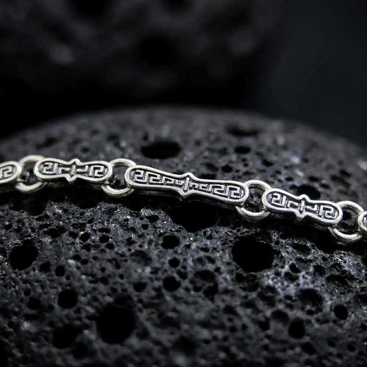 Oxidized Greek key chain necklace rests on dark volcanic rock in a macro studio close-up, showing detailed meander links with antique silver contrast.