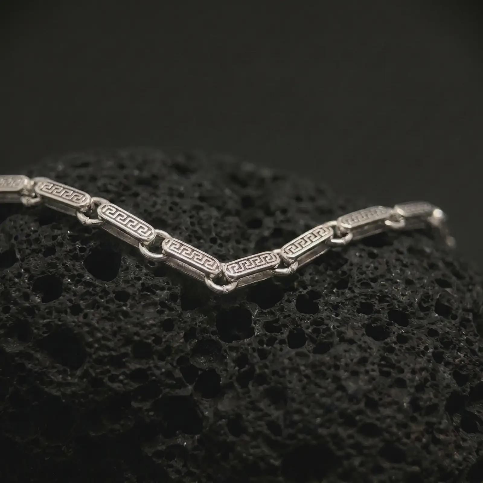 Polished silver Greek key chain necklace displayed on dark volcanic stone, with bright edges and detailed repeating geometric links clearly visible.