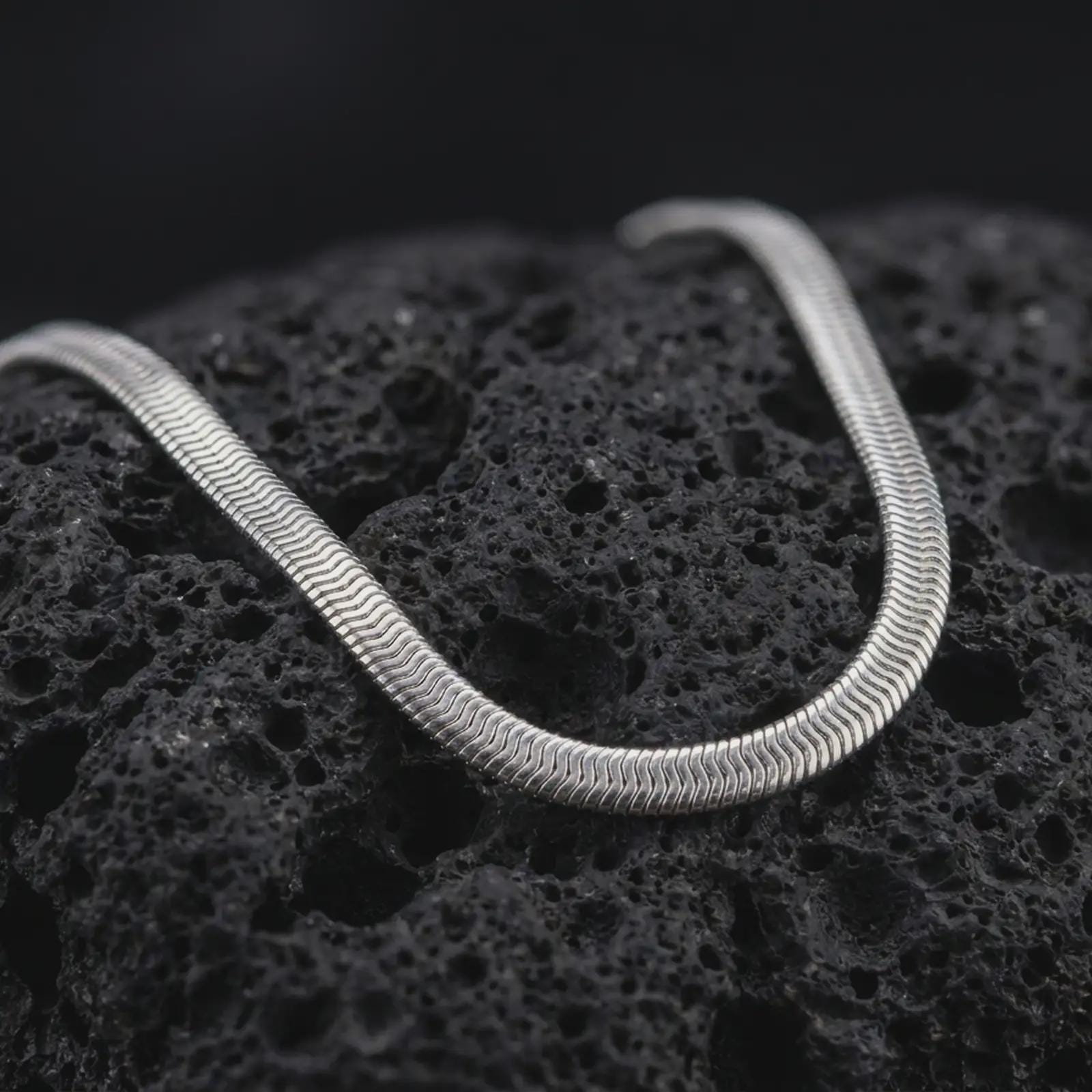 Polished silver snake chain necklace on volcanic stone, highlighting the sleek continuous surface and clean reflective finish.