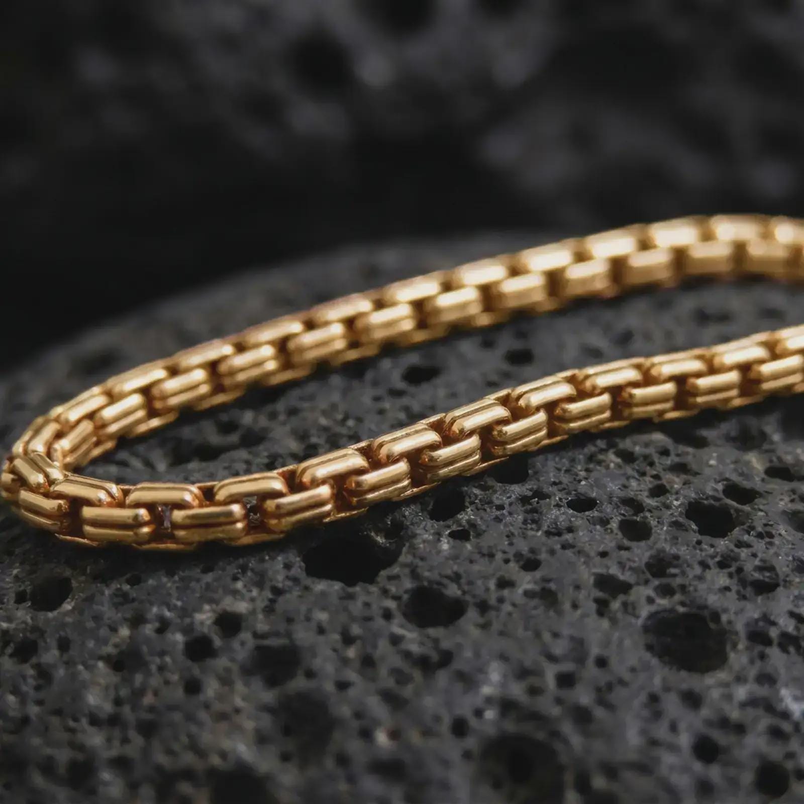 Gold plated box chain arranged across black volcanic rock, emphasizing its squared link construction and bold linear texture.