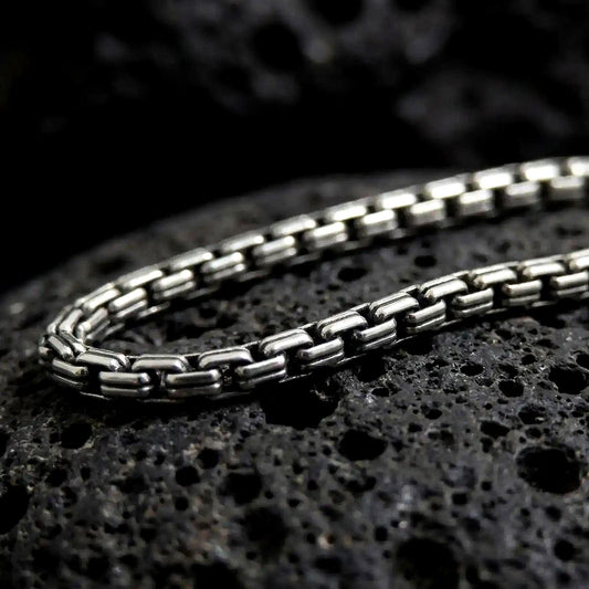 Oxidized sterling silver box chain displayed on black volcanic rock, showing its compact rectangular links and structured geometric profile.