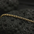 Load image into Gallery viewer, Gold plated Venetian box chain arranged across black volcanic rock, emphasizing its structured link pattern and refined metal surface.

