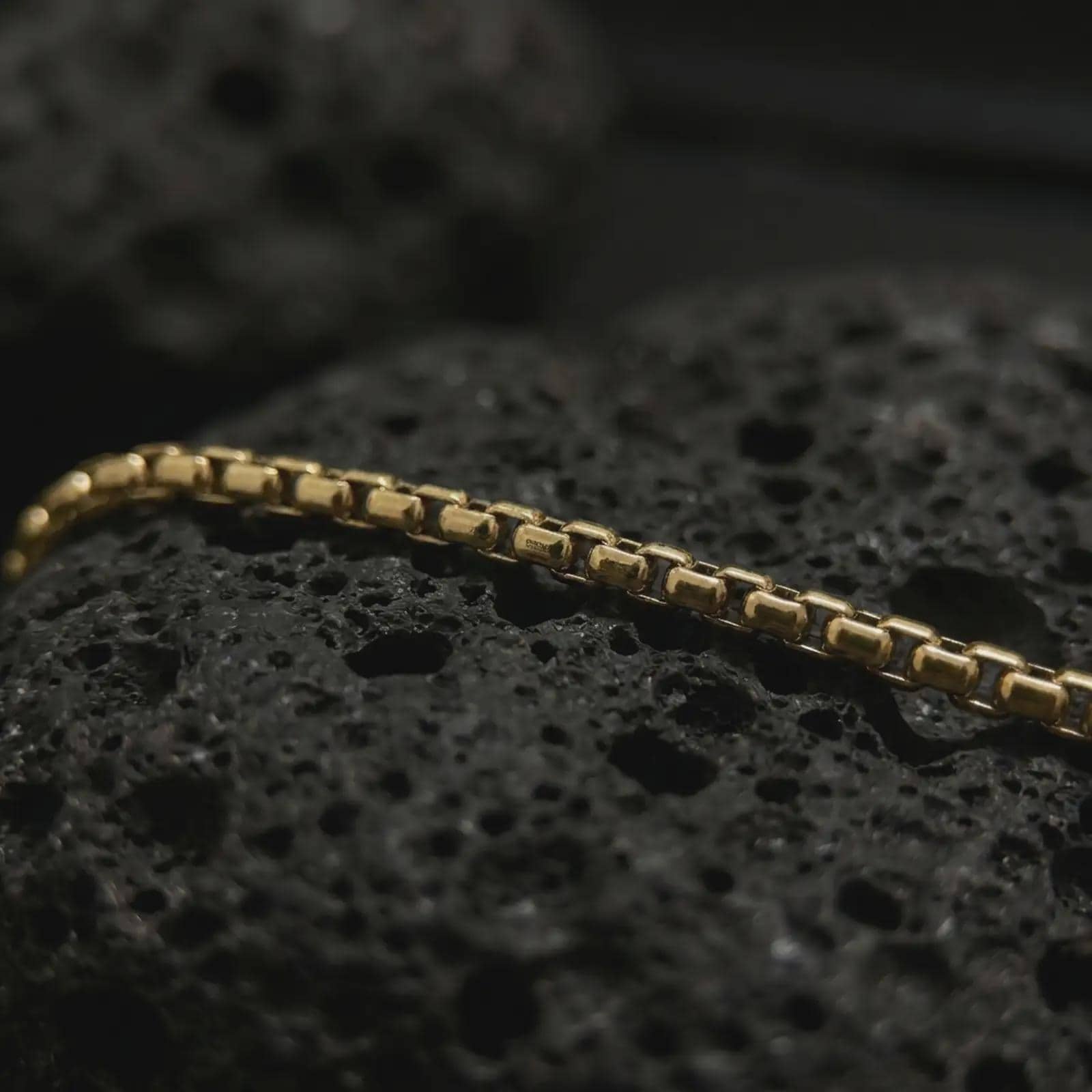 Gold plated Venetian box chain arranged across black volcanic rock, emphasizing its structured link pattern and refined metal surface.
