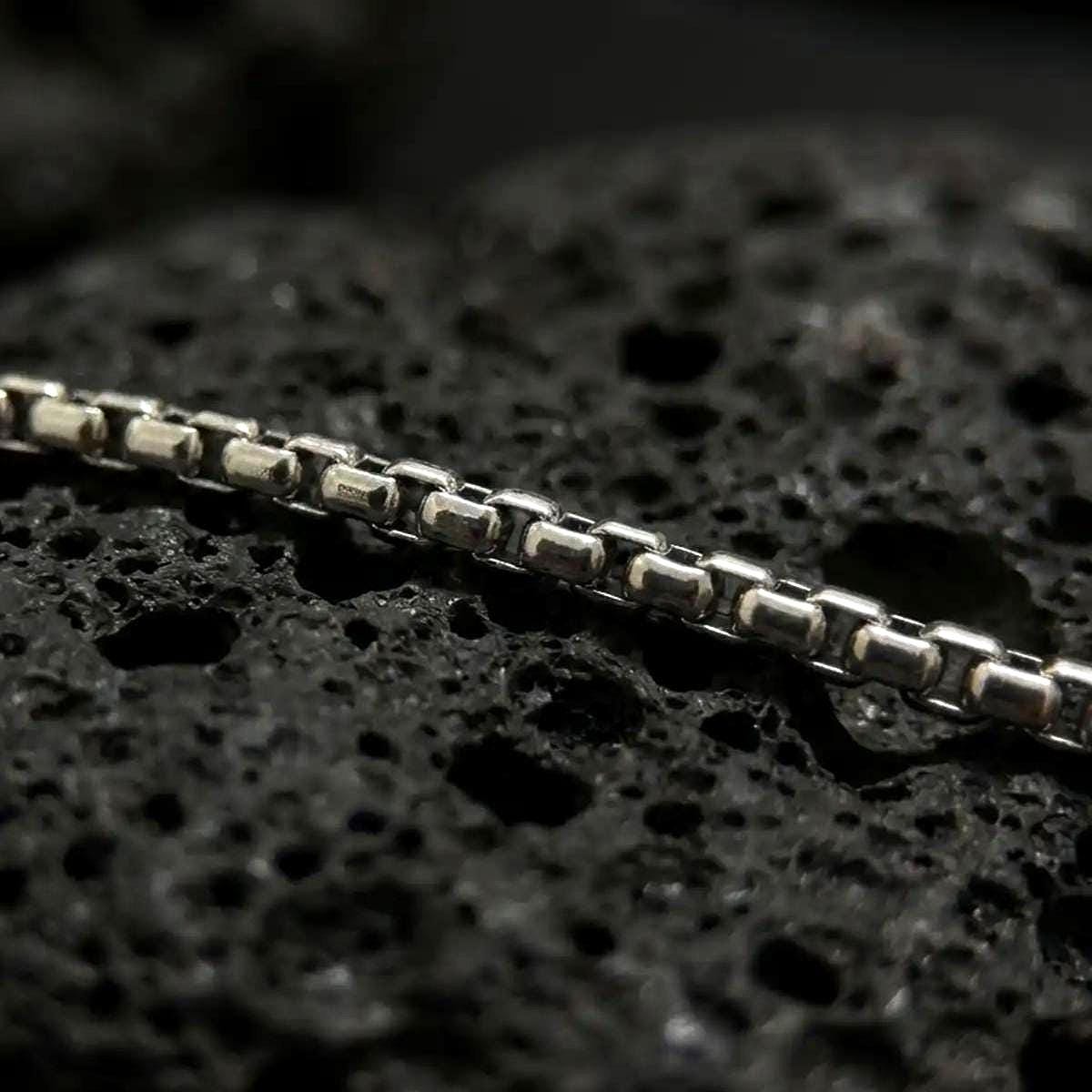 Oxidized sterling silver Venetian box chain displayed on black volcanic rock, showing its squared link structure and darkened detail.