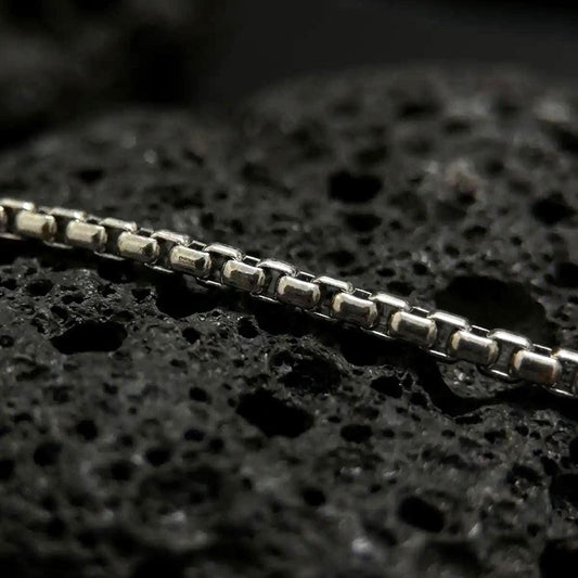 Oxidized sterling silver Venetian box chain displayed on black volcanic rock, showing its squared link structure and darkened detail.