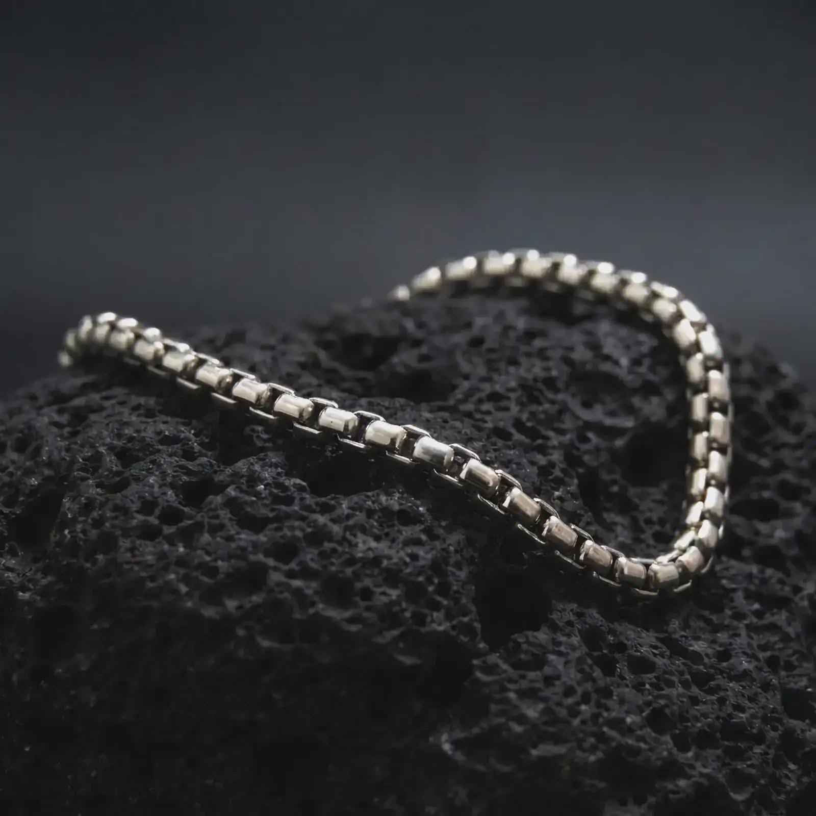 Polished silver box chain bracelet arranged across dark volcanic stone, highlighting the clean square-link structure and smooth reflective finish.