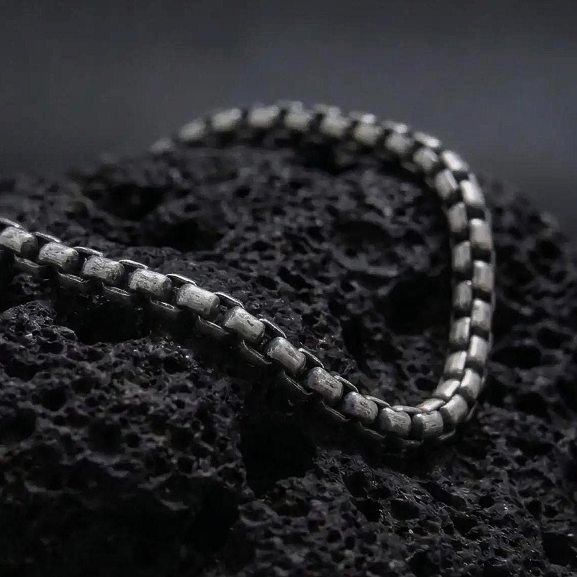 Oxidized sterling silver box chain bracelet displayed on black volcanic rock, showing its slim rectangular links and aged silver texture.