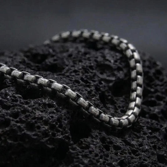 Oxidized sterling silver box chain bracelet displayed on black volcanic rock, showing its slim rectangular links and aged silver texture.