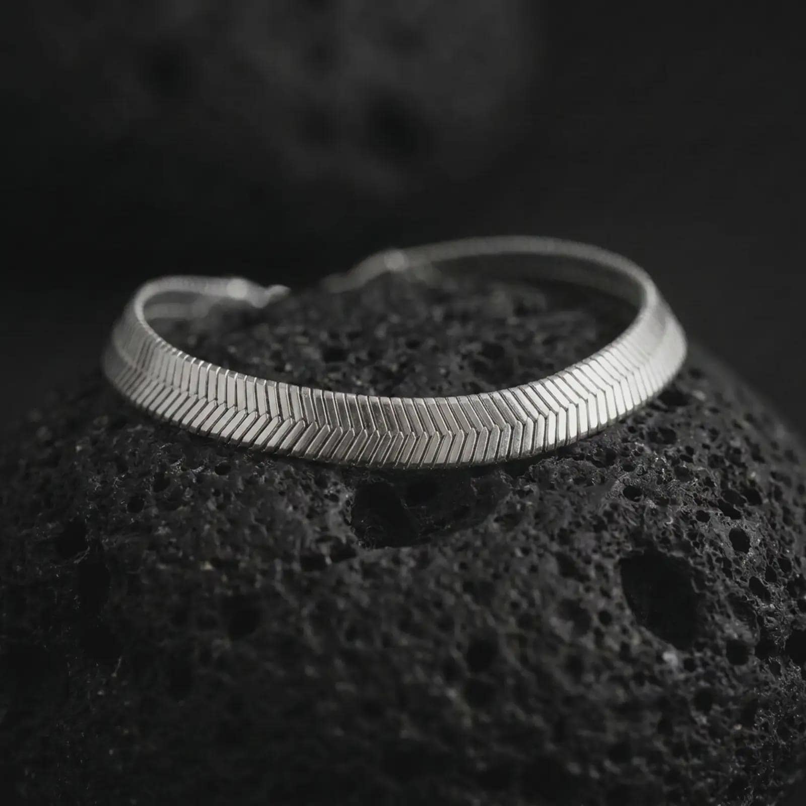 Polished silver herringbone chain bracelet arranged on dark volcanic stone with a clean reflective surface and precise link pattern.
