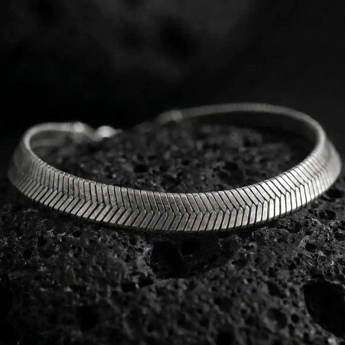 Oxidized sterling silver herringbone bracelet displayed on black volcanic rock, showing its slim flat profile and chevron texture.