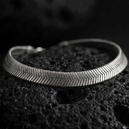 Oxidized sterling silver herringbone bracelet displayed on black volcanic rock, showing its slim flat profile and chevron texture.