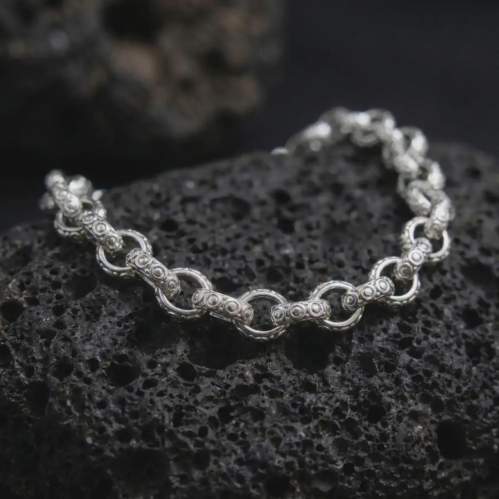 Polished silver ornamental bracelet arranged on dark volcanic stone, featuring rounded connector links and decorative bar-shaped elements.