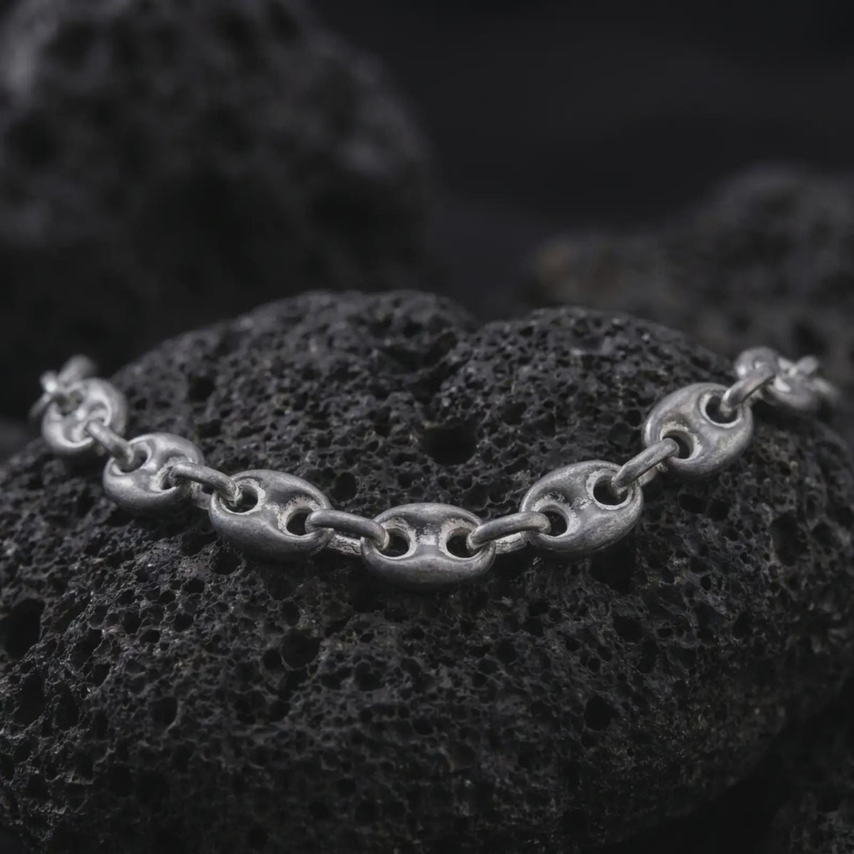 An 11mm mariner chain necklace with an oxidized finish rests on dark volcanic rock, showing substantial oval anchor links with recessed central bars in a macro studio close-up.