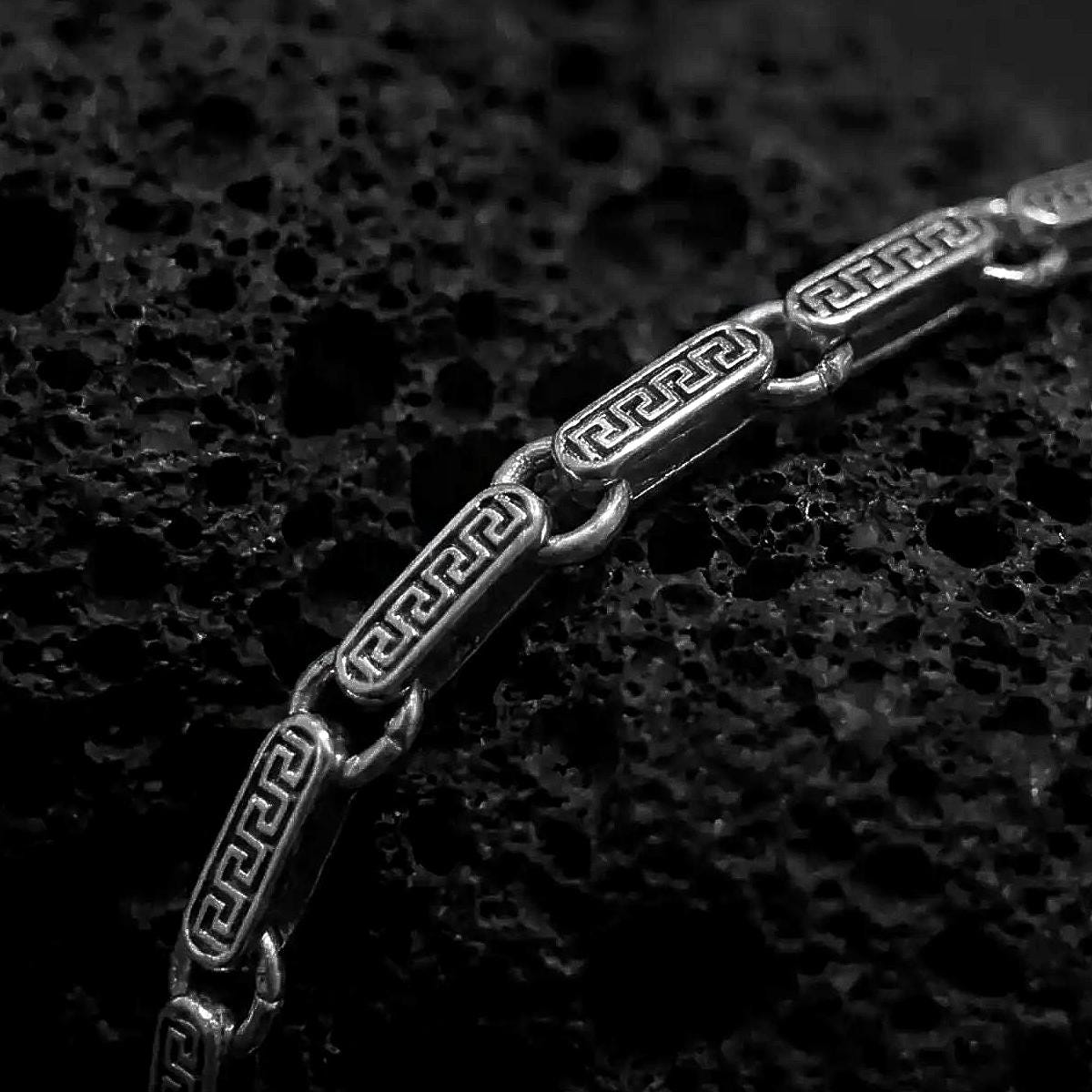 Greek key bracelet with an oxidized finish displayed across dark volcanic rock in a premium macro studio close-up.