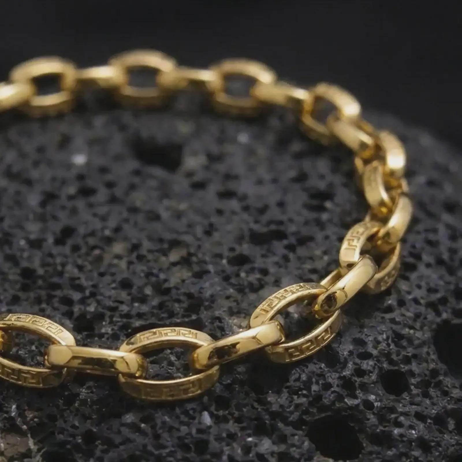 Gold plated Greek key bracelet resting on rugged volcanic rock, with detailed eternity motifs engraved around each circular link in a luxury macro studio close-up.