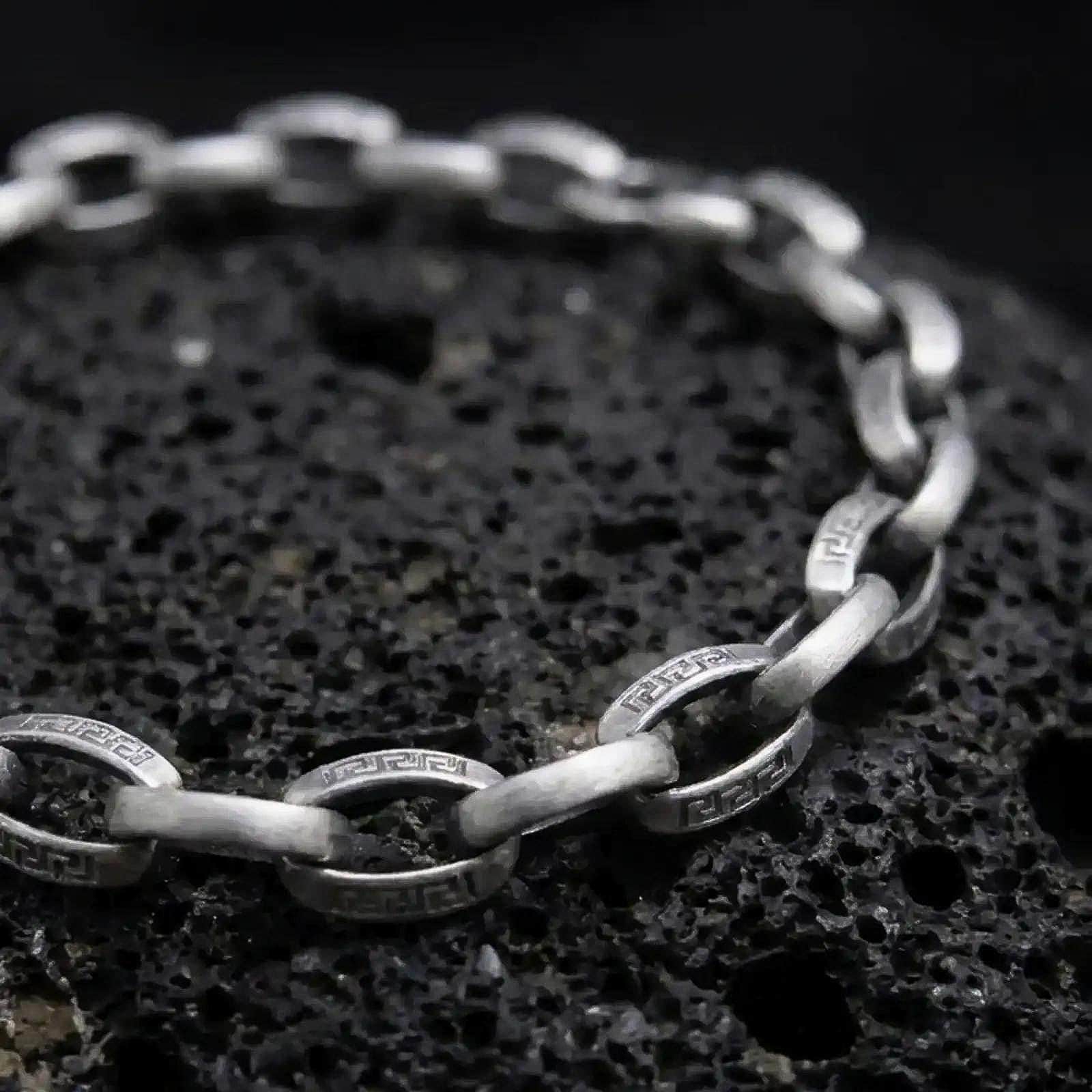 Sterling silver Greek key bracelet with an oxidized finish arranged on dark volcanic rock, showing engraved circular meander links and smooth rectangular connectors in a macro studio close-up.