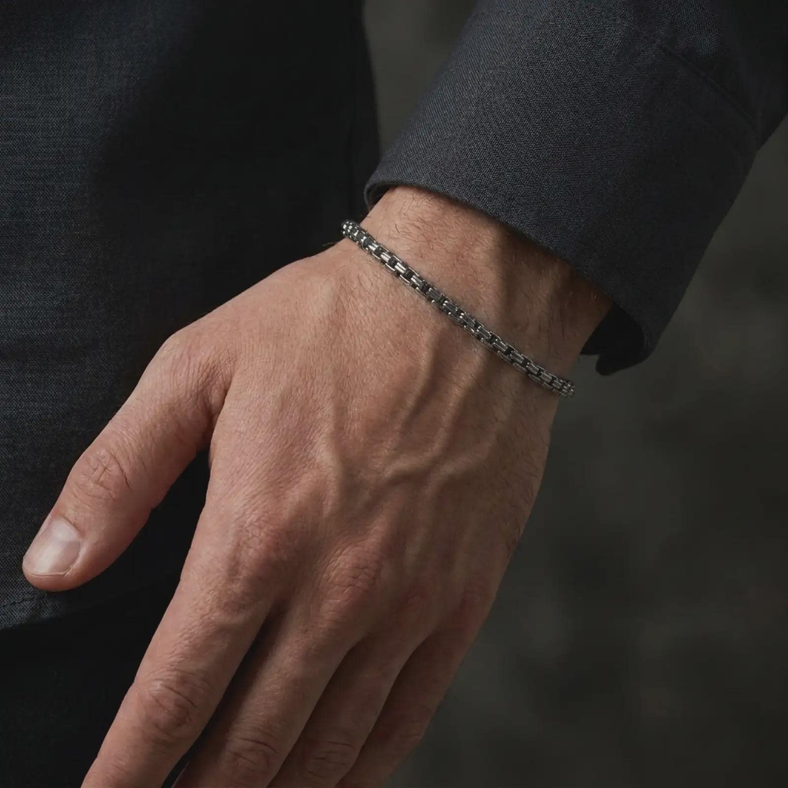 Men’s sterling silver box chain bracelet with an oxidized finish worn closely around the wrist, showing its slim geometric structure against dark formal clothing in a masculine lifestyle close-up.