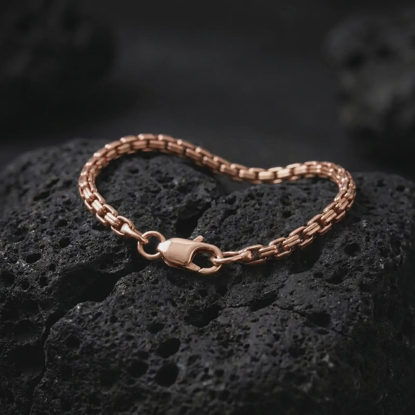 Rose gold plated slim box chain bracelet arranged on rugged black volcanic rock, emphasizing the structured interlocking links and minimalist profile in a refined macro studio image.