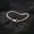 Load image into Gallery viewer, Polished silver box chain bracelet displayed on textured volcanic rock, highlighting the closely connected rectangular links, flexible construction, and secure clasp in a premium macro photograph.
