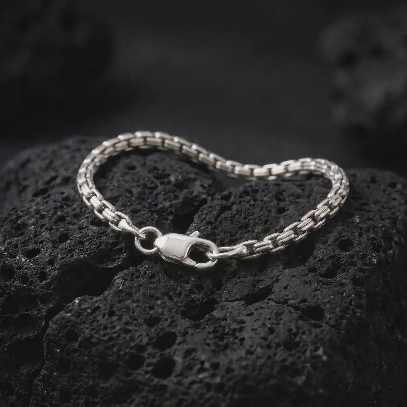 Polished silver box chain bracelet displayed on textured volcanic rock, highlighting the closely connected rectangular links, flexible construction, and secure clasp in a premium macro photograph.