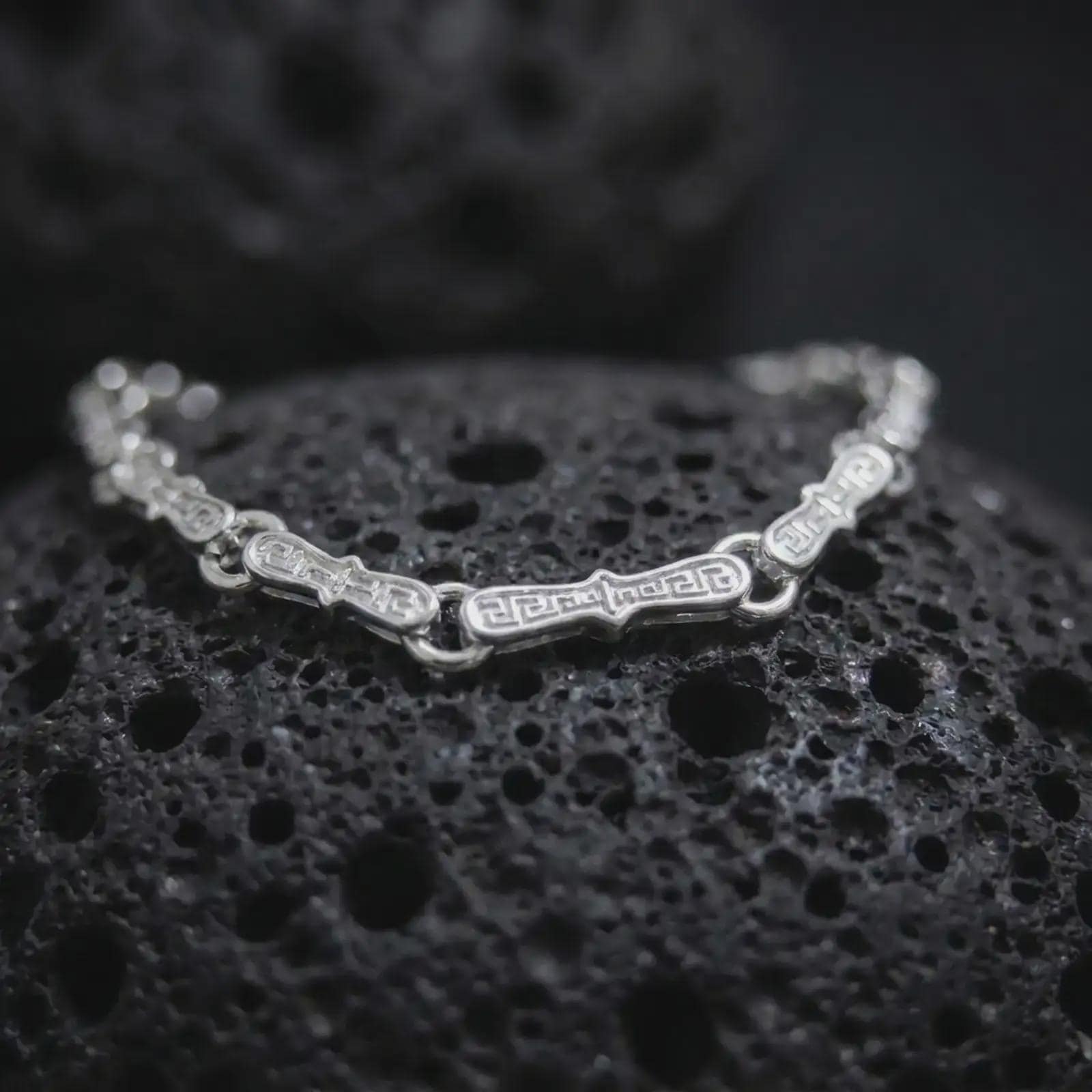 Polished silver Greek meander bracelet displayed across textured black volcanic rock, highlighting the sculpted geometric links and continuous recessed pattern in a premium macro studio photograph.