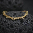 Load image into Gallery viewer, Gold plated Greek key link bracelet resting on rugged volcanic rock, featuring elongated symbolic links connected by smooth round rings in a luxury macro studio close-up.
