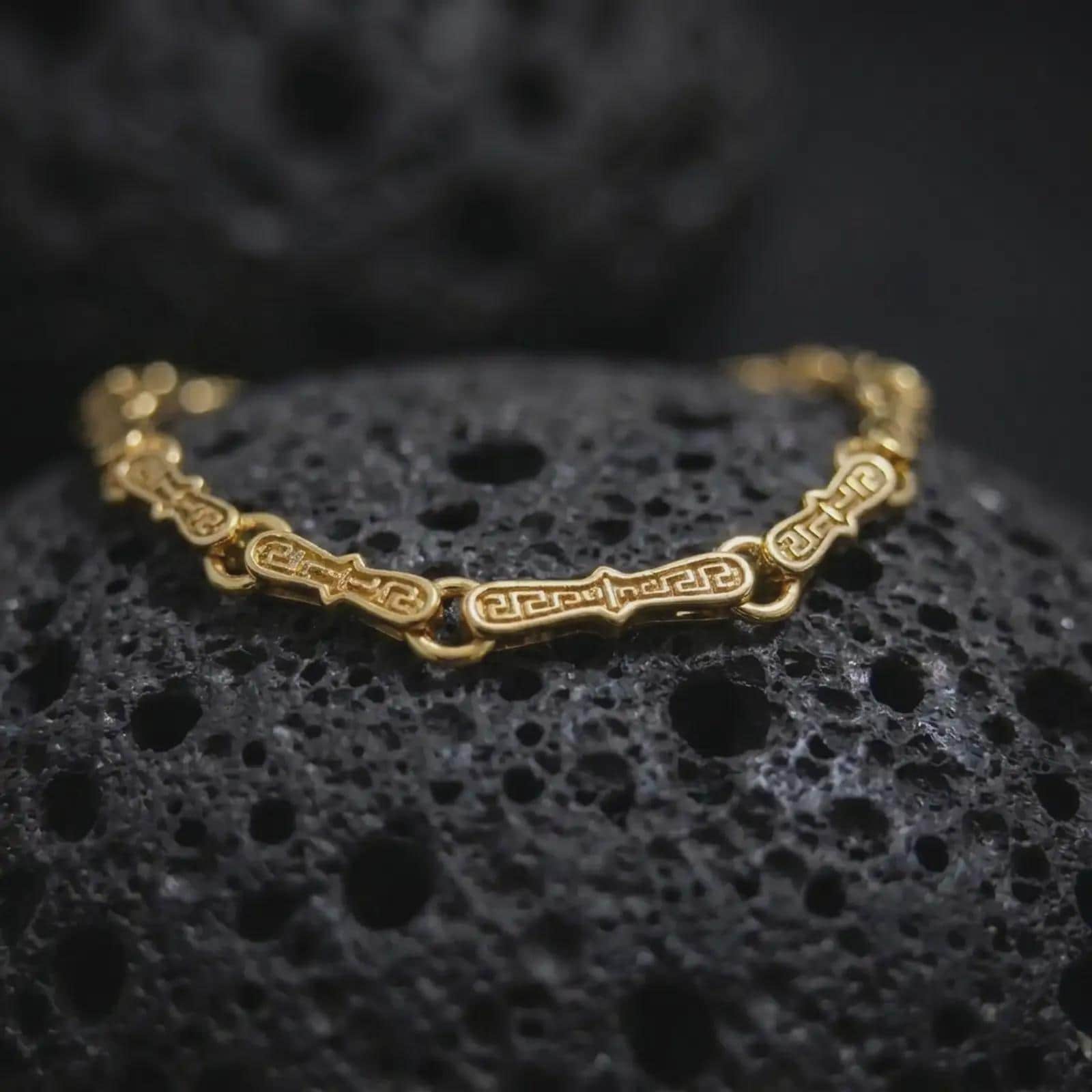 Gold plated Greek key link bracelet resting on rugged volcanic rock, featuring elongated symbolic links connected by smooth round rings in a luxury macro studio close-up.