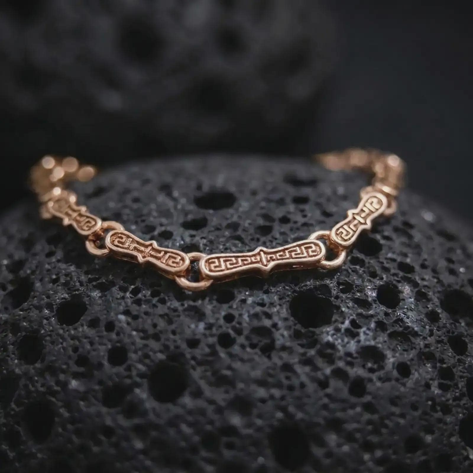 Rose gold plated Greek meander bracelet arranged on black volcanic rock, emphasizing the repeating eternity pattern and articulated link construction in a refined macro photograph.
