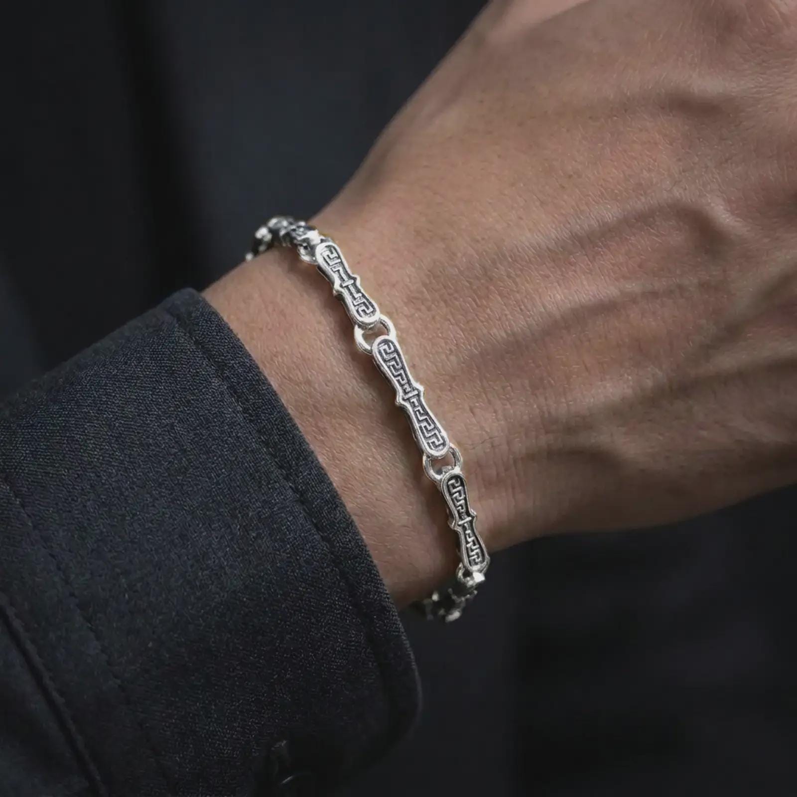 Polished silver Greek meander link bracelet worn around a man’s wrist, showing its articulated circular connectors and refined geometric profile in a detailed lifestyle close-up.