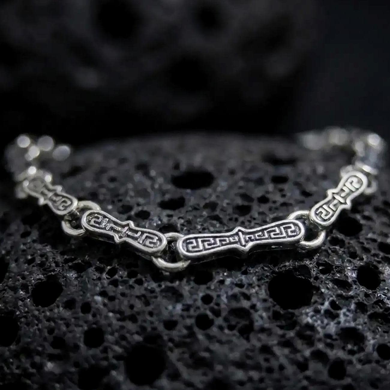 Oxidized sterling silver Greek key bracelet arranged on dark volcanic rock, showing elongated engraved meander links and circular connectors in a detailed macro studio close-up.
