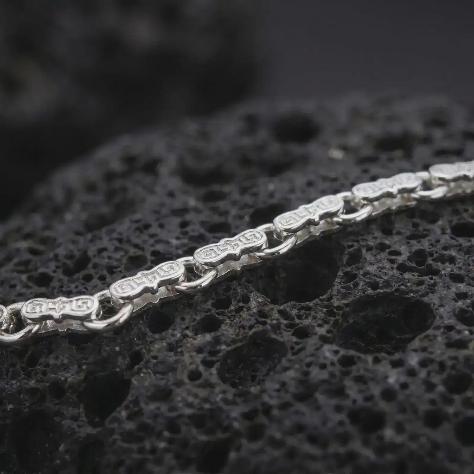 The polished silver Greek meander bracelet is arranged on textured black volcanic rock, highlighting its double-ended bar links and smooth circular connectors in a premium macro photograph.