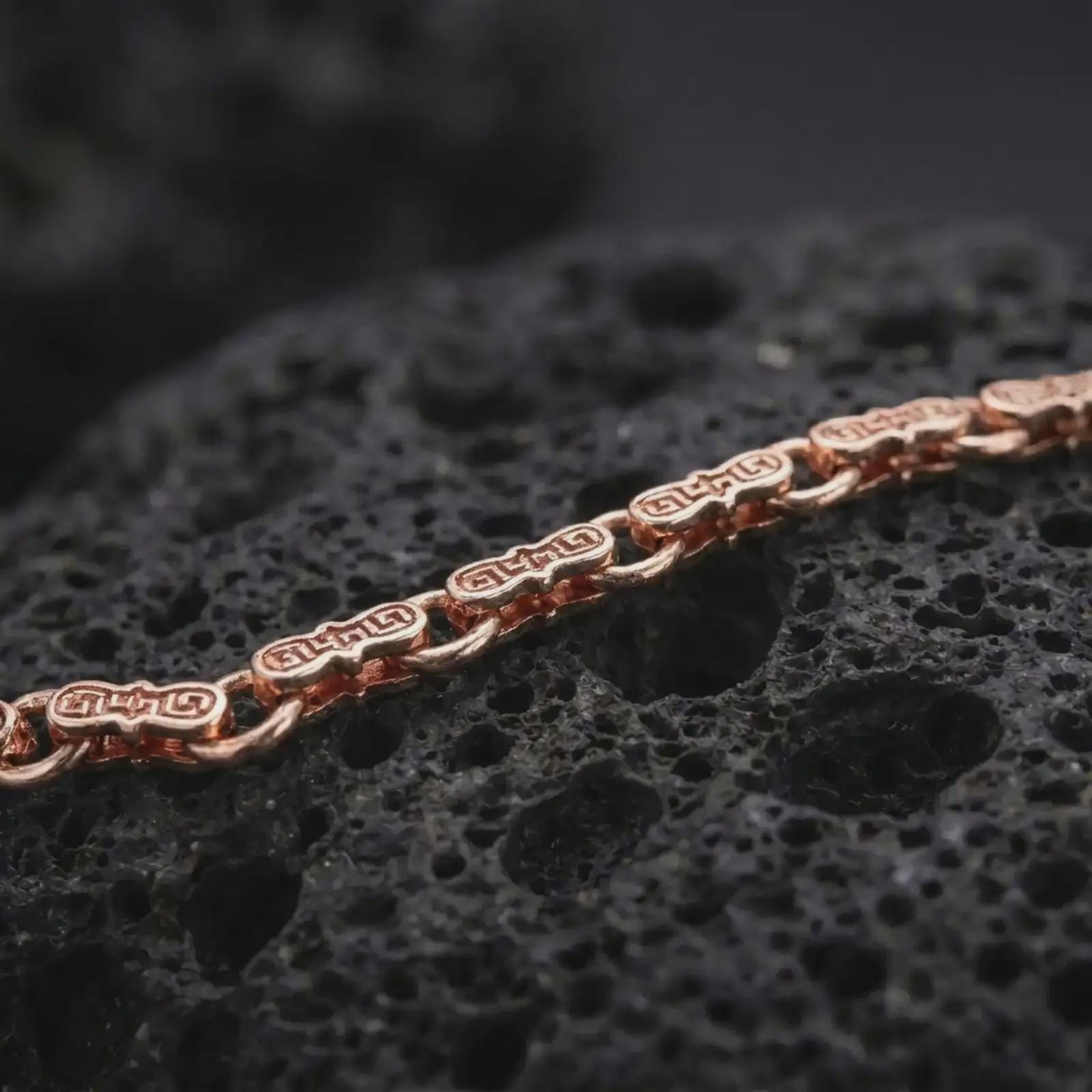The rose gold plated meander bracelet is displayed across black volcanic rock, emphasizing its slim profile, sculpted bar links, and ancient Greek detailing in a refined macro image.