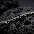 Load image into Gallery viewer, A Greek key bracelet with an oxidized finish rests across dark volcanic rock, showing sculpted hourglass-shaped links and recessed meander engravings in a macro studio close-up.
