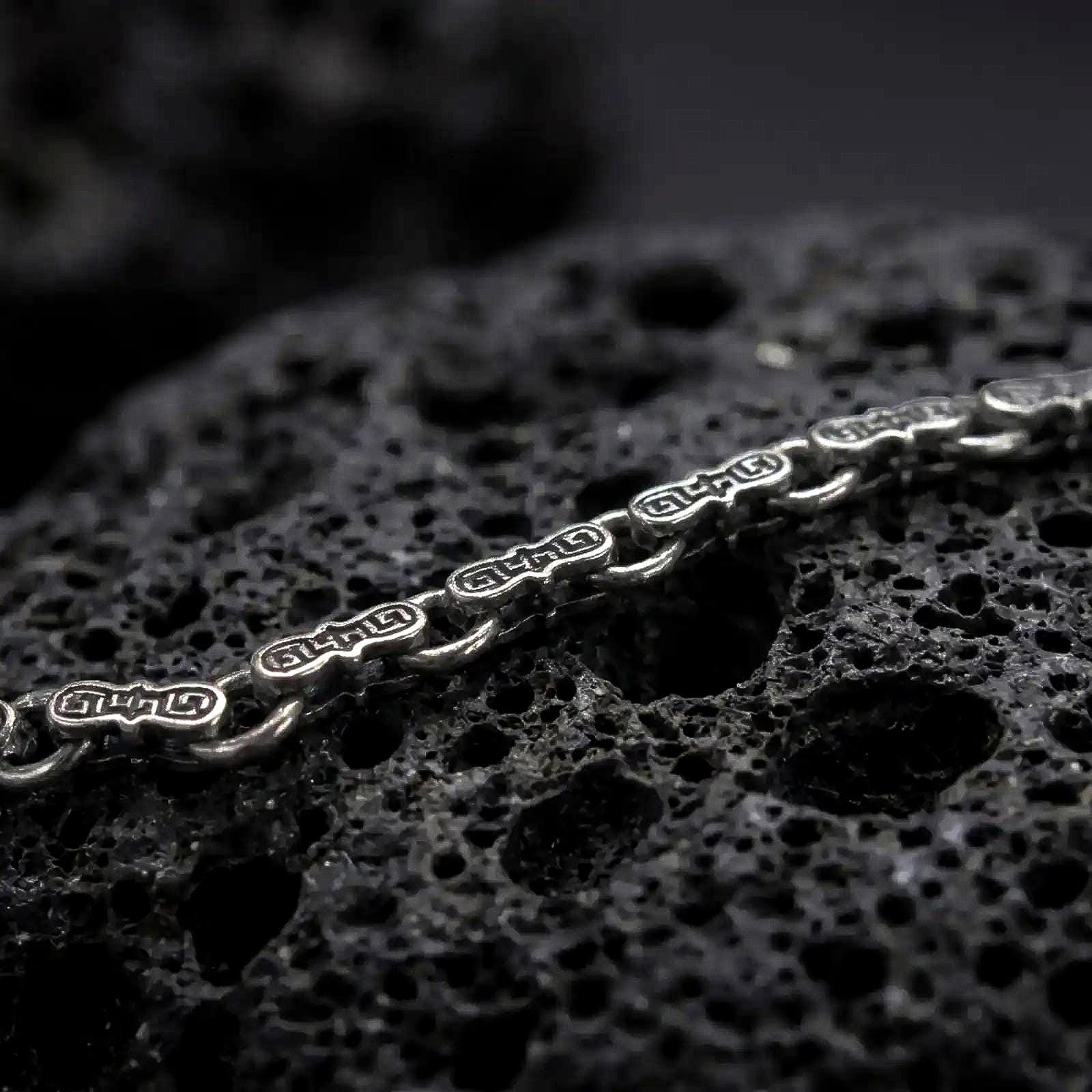A Greek key bracelet with an oxidized finish rests across dark volcanic rock, showing sculpted hourglass-shaped links and recessed meander engravings in a macro studio close-up.