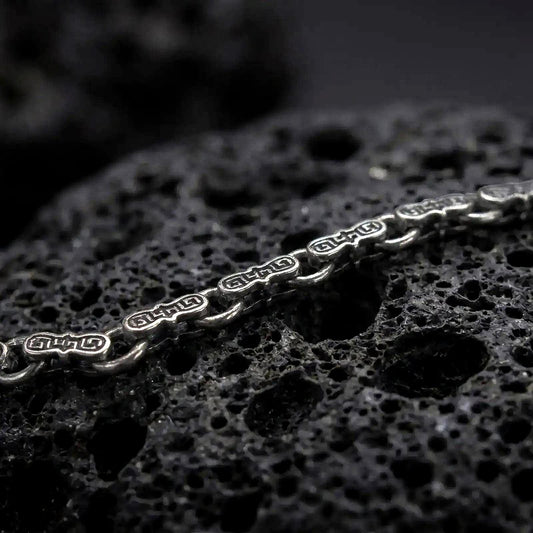 A Greek key bracelet with an oxidized finish rests across dark volcanic rock, showing sculpted hourglass-shaped links and recessed meander engravings in a macro studio close-up.