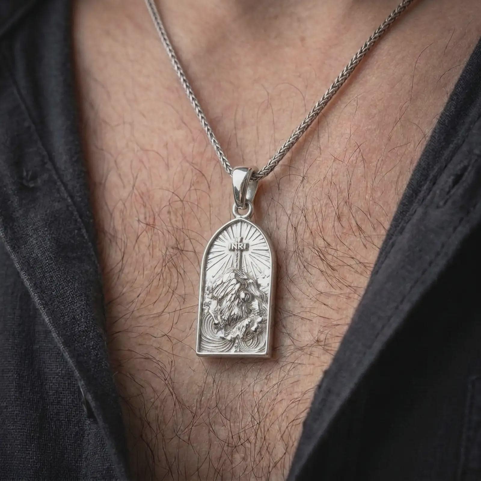 The polished silver Christian pendant is worn on a man’s chest, with Jesus beneath the cross, radiant background lines, and the arched medallion shape clearly visible in a premium lifestyle photograph.
