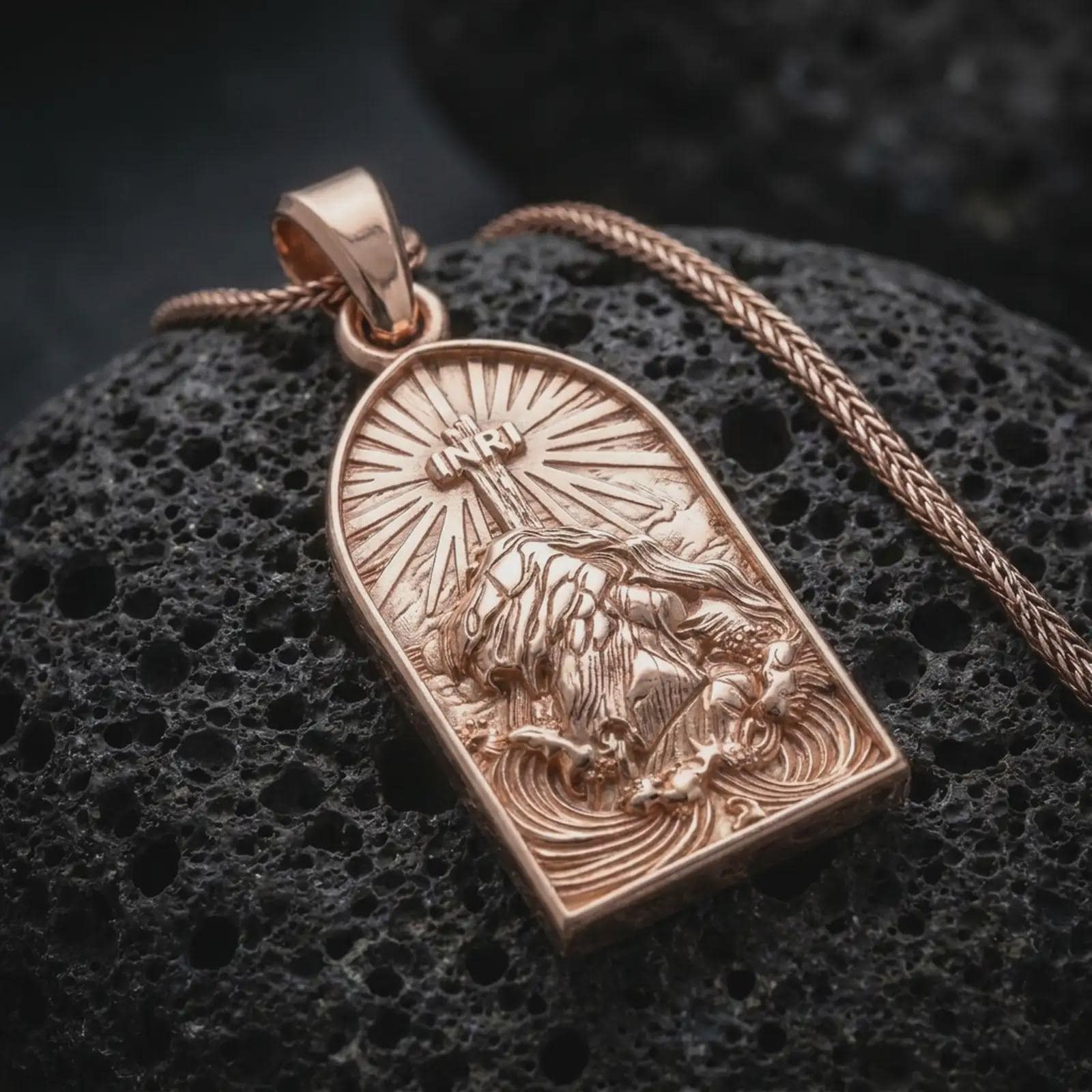 The rose gold plated INRI pendant is arranged on rugged volcanic rock, emphasizing the sculpted figure of Jesus, the cross above him, and the ornamental arched frame in a refined macro image.