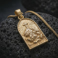 Load image into Gallery viewer, A gold plated Jesus carrying the cross pendant lies on black volcanic rock, featuring Christ kneeling beneath the cross with radiant lines and intricate devotional details in a luxury macro studio close-up.
