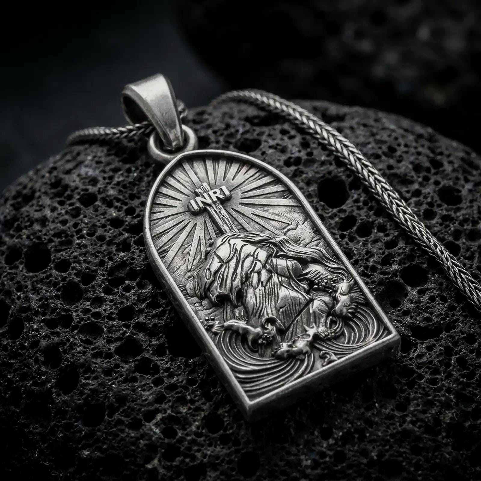 An oxidized Jesus carrying the cross pendant rests on dark volcanic rock, showing the INRI inscription, radiant background, kneeling figure, and sculpted religious details in a macro studio close-up.