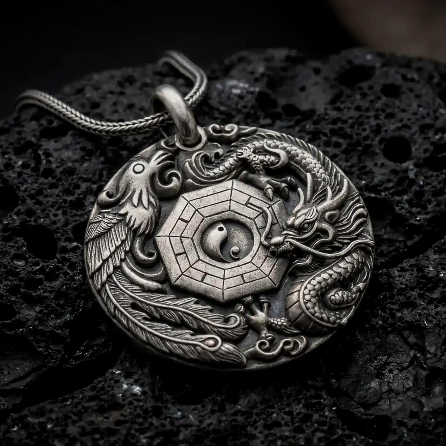 An oxidized dragon and phoenix pendant rests on dark volcanic rock, showing the central Yin Yang symbol, octagonal Bagua design, detailed scales, flowing feathers, and cloud motifs in a macro studio close-up.
