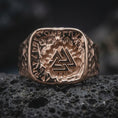 Load image into Gallery viewer, The rose gold plated Viking signet ring is arranged on black volcanic rock, emphasizing its substantial square face, interlocking triangular emblem, engraved rune border, and textured side design in a refined macro image.
