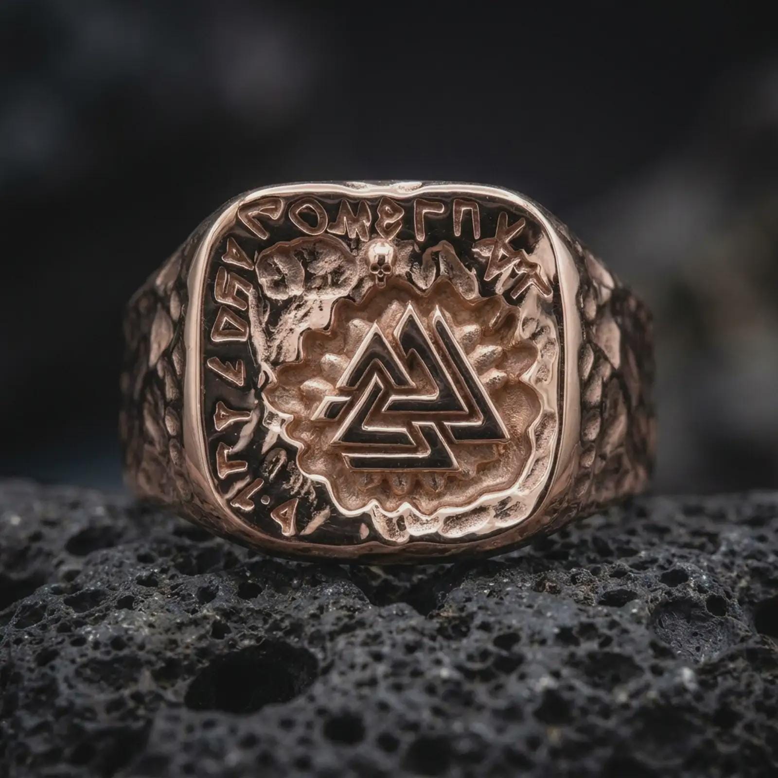 The rose gold plated Viking signet ring is arranged on black volcanic rock, emphasizing its substantial square face, interlocking triangular emblem, engraved rune border, and textured side design in a refined macro image.