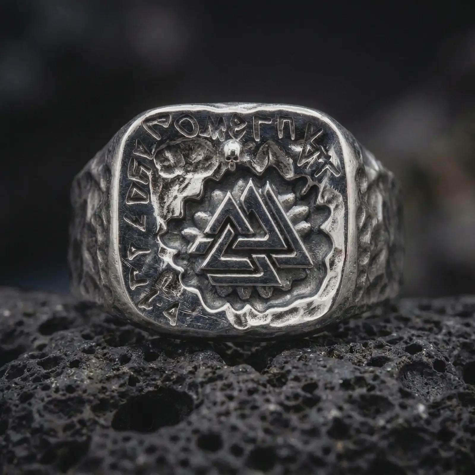 The polished silver Viking signet ring is displayed on textured black volcanic rock, highlighting the central Valknut emblem, rune border, sculpted skull, and detailed masculine shoulders in a premium macro photograph.