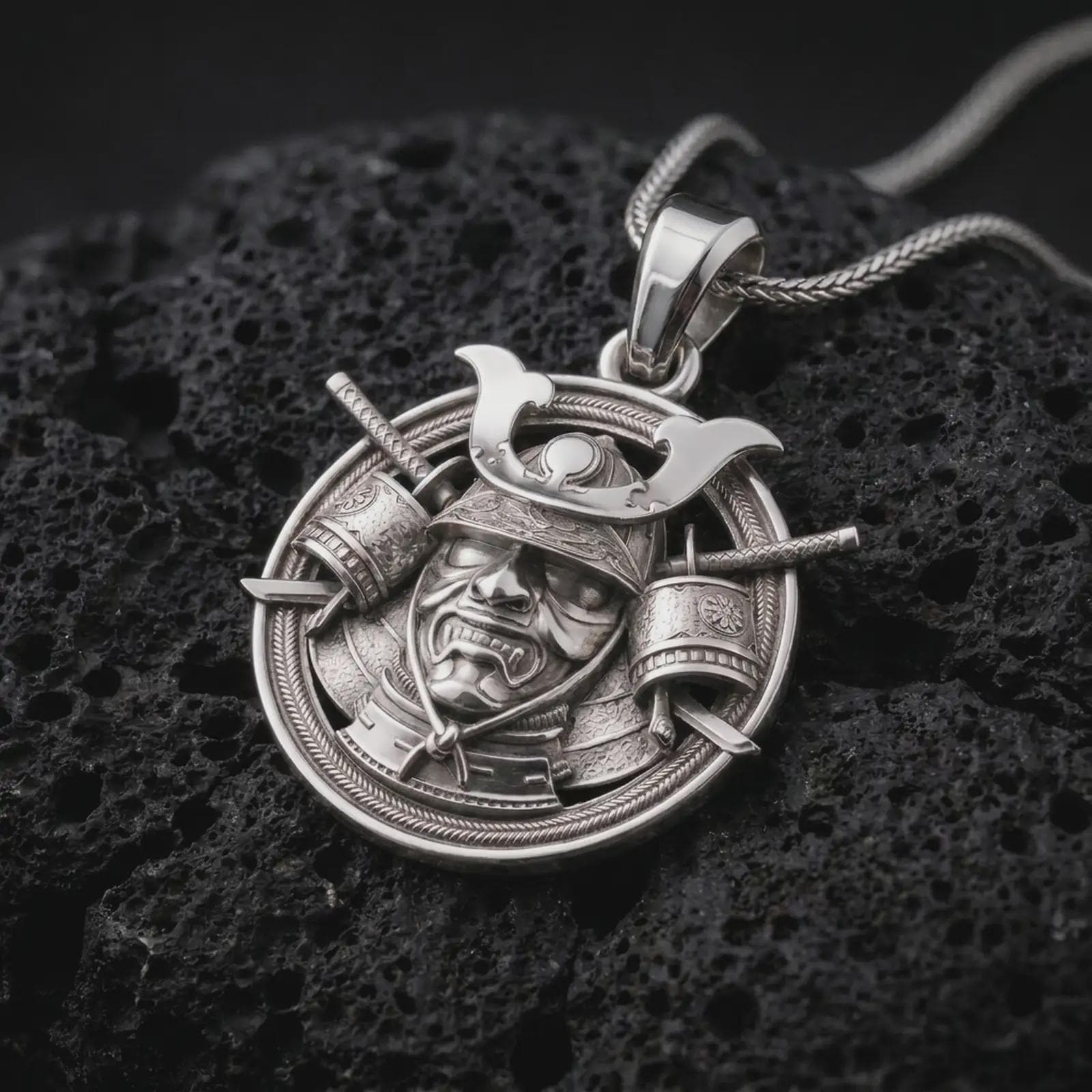 The polished silver samurai pendant is displayed on textured volcanic rock, highlighting the sculpted warrior face, engraved helmet crest, layered armor, and dimensional Japanese details in a premium macro photograph.